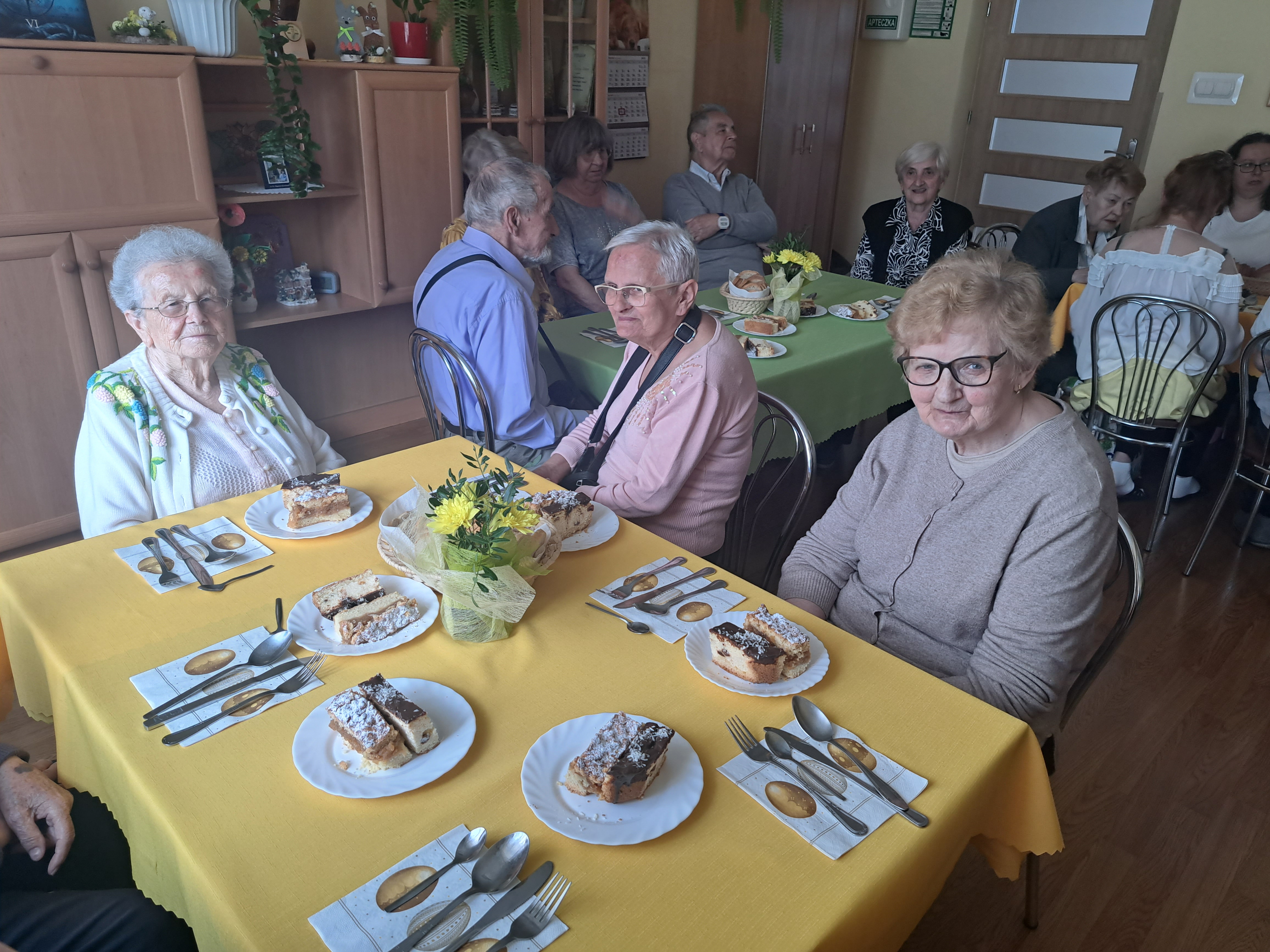 Trzy Seniorki siedzą przed zastawionym stołem, na talerzykach leżą ciasta